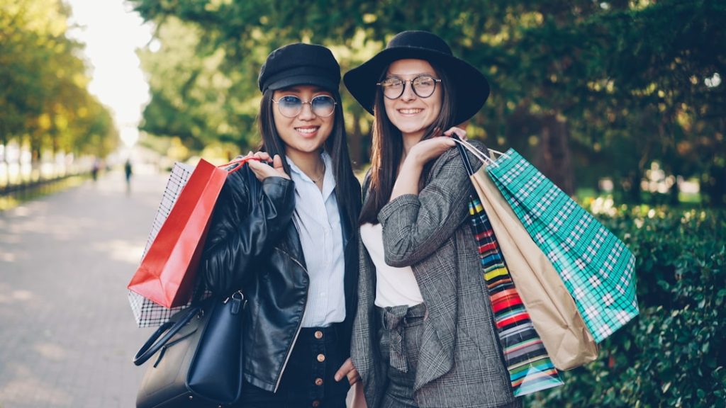 two-smiling-women-holding-shopping-bags-outdoors-wvobj0ujhsi Portrait of beautiful young women shoppers standing in the street holding paper bags and smiling looking at camera. Youth lifestyle, friendship and consumerism concept.