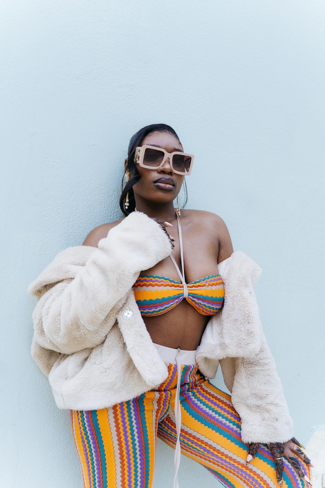 A stylish woman posing confidently against a clean minimal background, wearing bold oversized sunglasses, a soft beige faux-fur jacket, and colorful striped pants. The image blends fashion, color, texture, and modern editorial aesthetics.